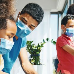 Family of four, wearing face masks, looking through a window while under home isolation/quarantine during COVID-19 pandemic.